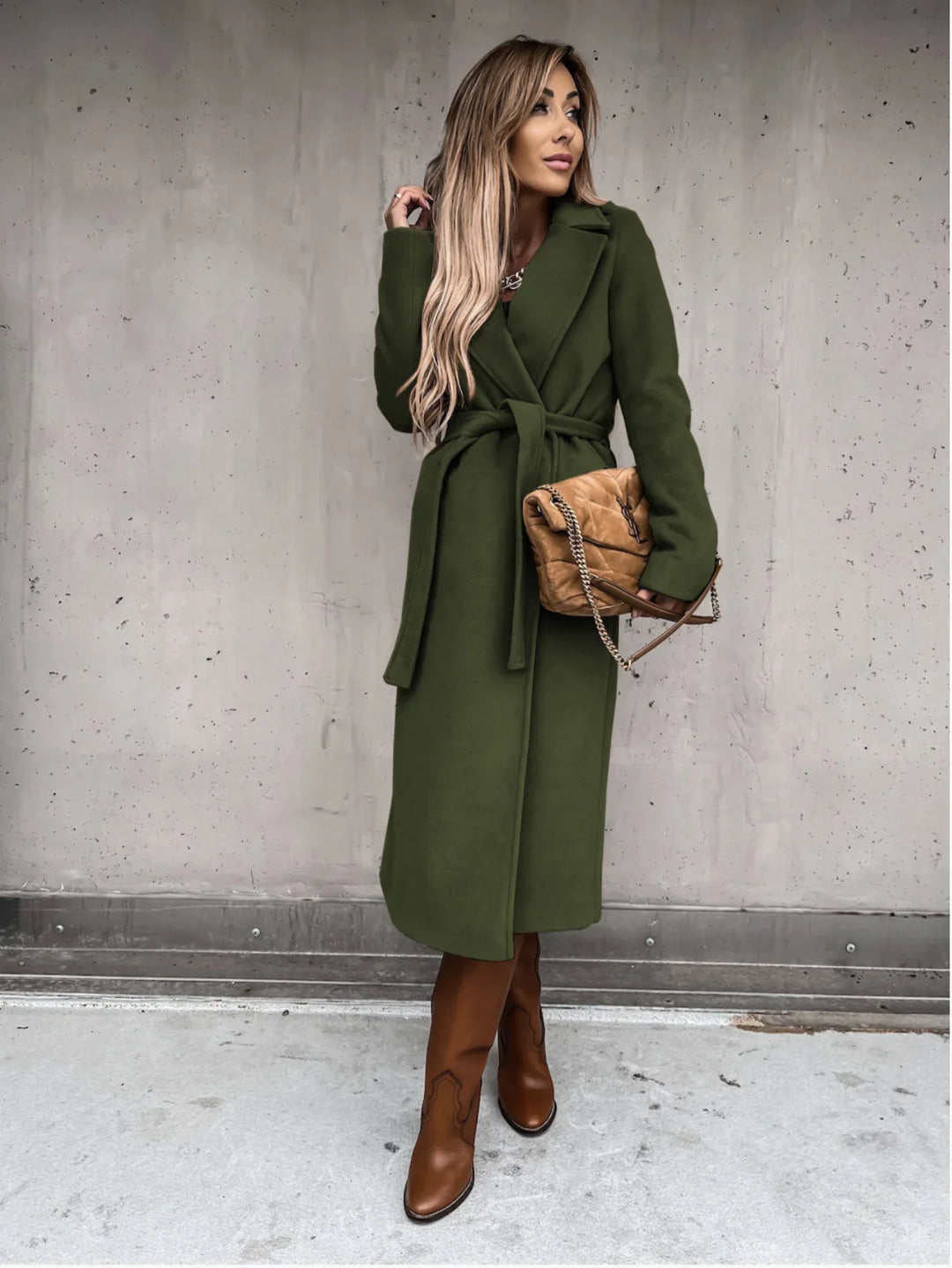 Woman in long olive green belted coat with tan leather boots and brown quilted handbag against concrete wall
