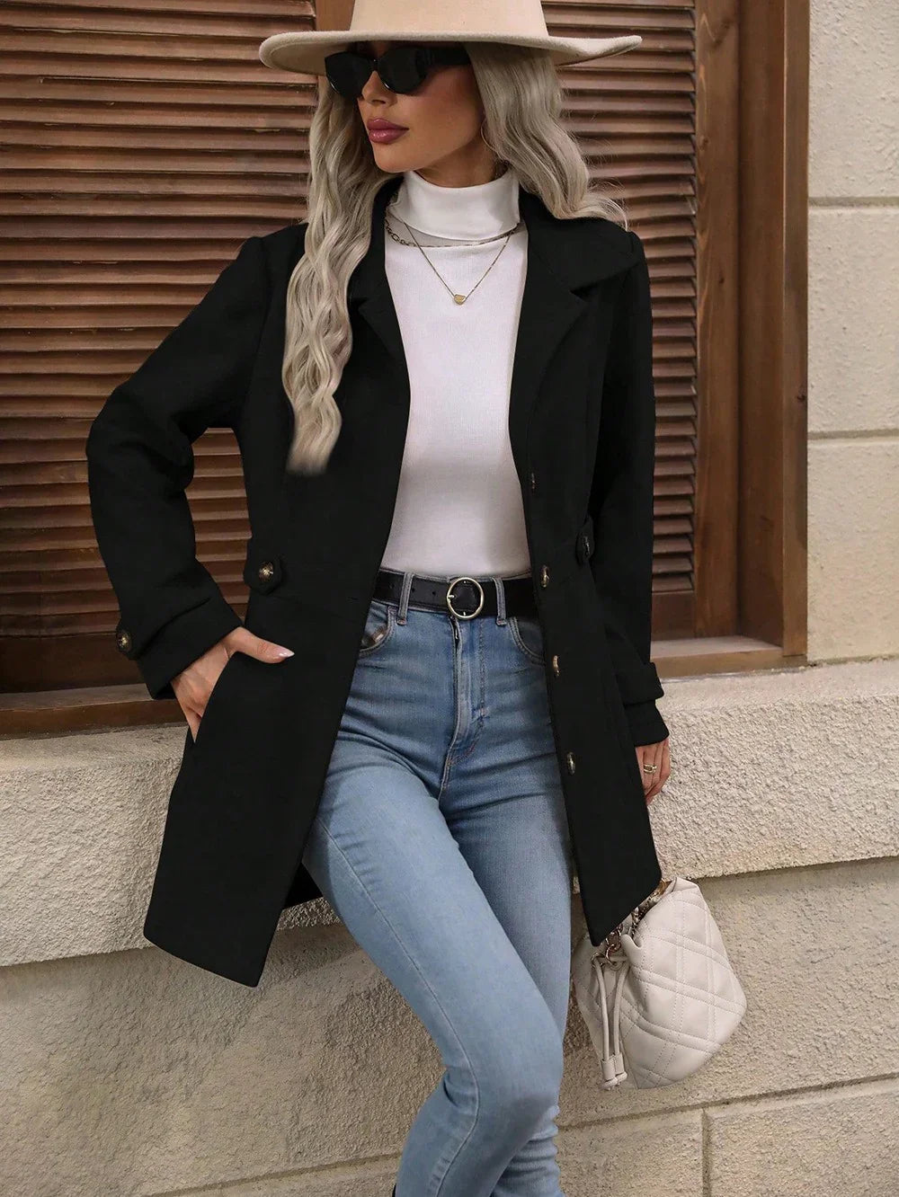 Woman in black coat, white turtleneck, blue jeans, beige hat, sunglasses, and quilted white handbag