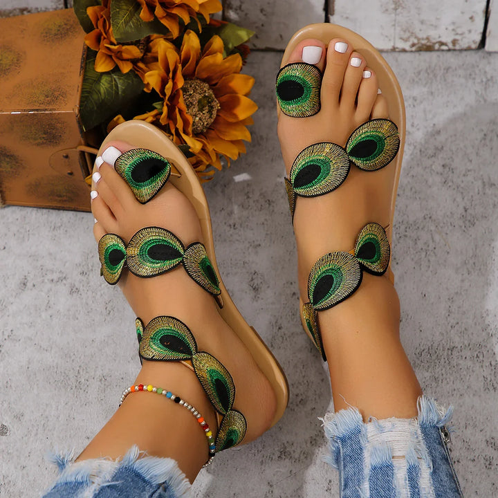 Feet wearing beige sandals with green peacock feather design straps on gray stone floor