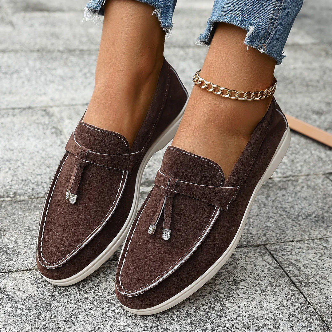 Brown suede loafers with white stitching and tassels worn with ripped blue jeans and gold ankle bracelet