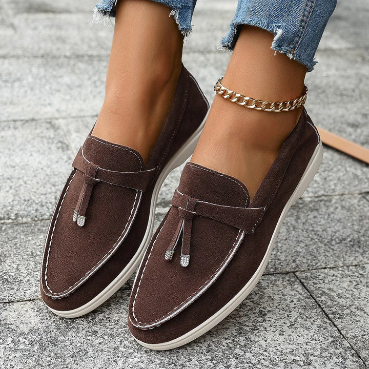 Brown suede loafers with white stitching and tassels worn with ripped blue jeans and gold ankle bracelet