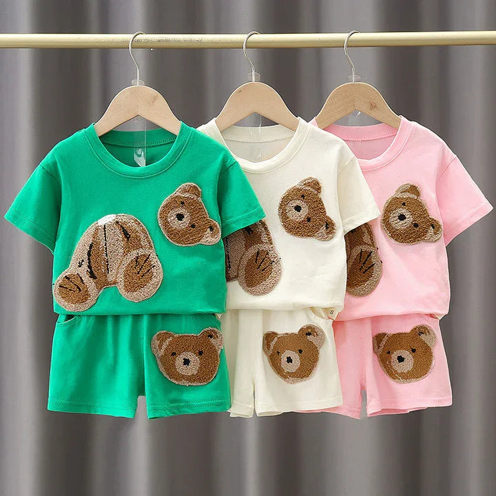 Three kids' short-sleeve outfits with teddy bear patches in green, white, and pink hanging on wooden hangers