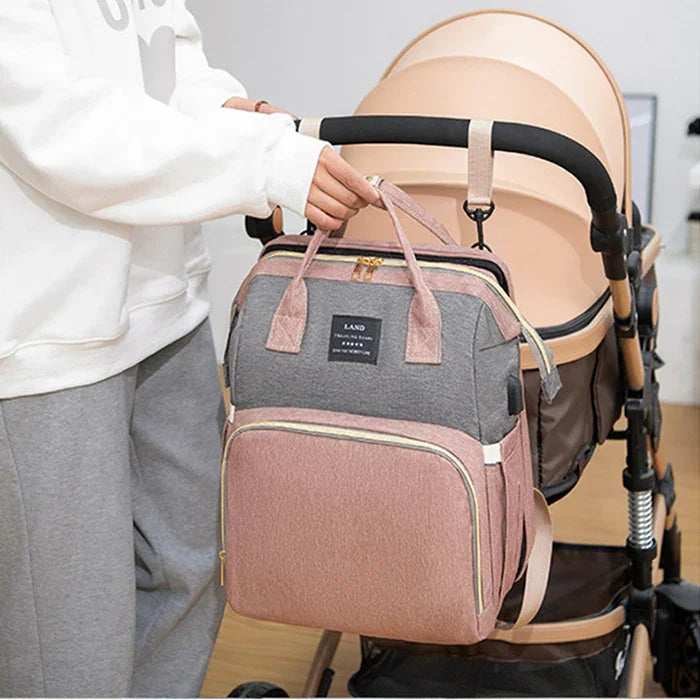 Person holding pink and gray diaper bag attached to tan baby stroller in indoor setting