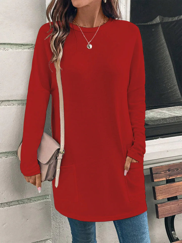 Woman wearing casual red long sleeve tunic top with front pockets and beige crossbody bag, standing near white brick wall