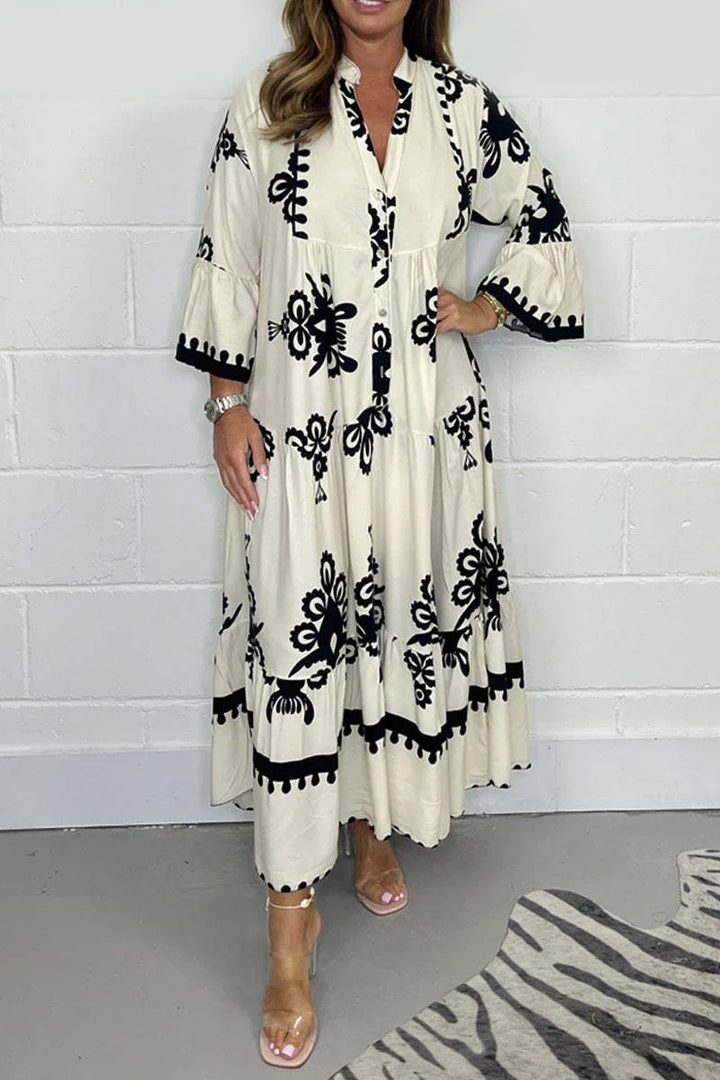 Woman wearing long beige dress with black floral embroidery and clear heels against white wall