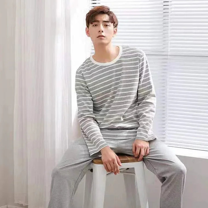 Young man wearing gray striped long-sleeve top and gray pants sitting on stool by window blinds