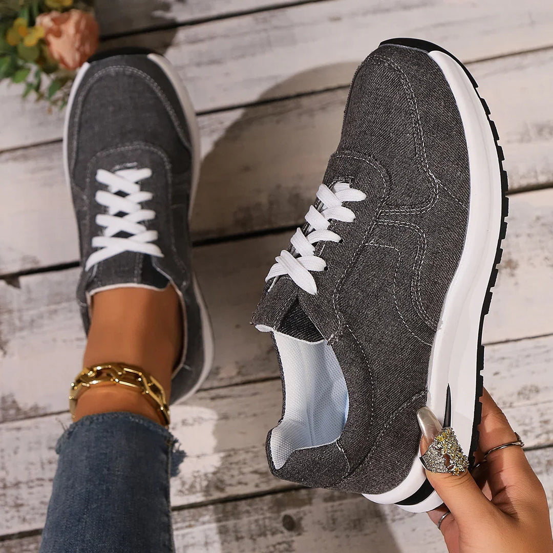 Casual dark gray sneakers with white laces worn on feet, styled with jeans and gold anklet on wooden floor
