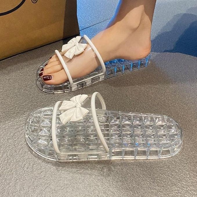 Transparent slide sandals with white straps and bow detail on textured flooring, one worn on foot