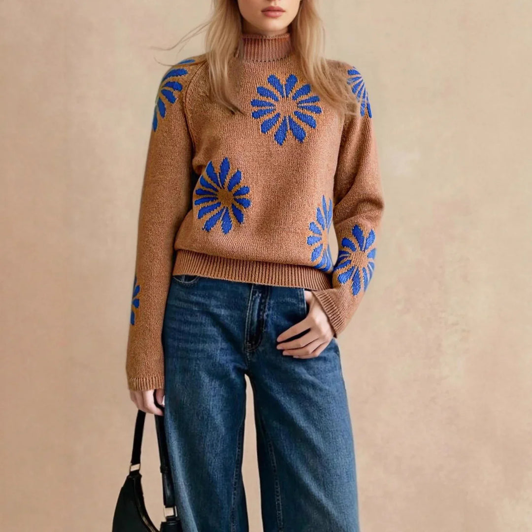 Woman wearing brown knit sweater with blue floral patterns and high-waisted blue jeans holding black handbag