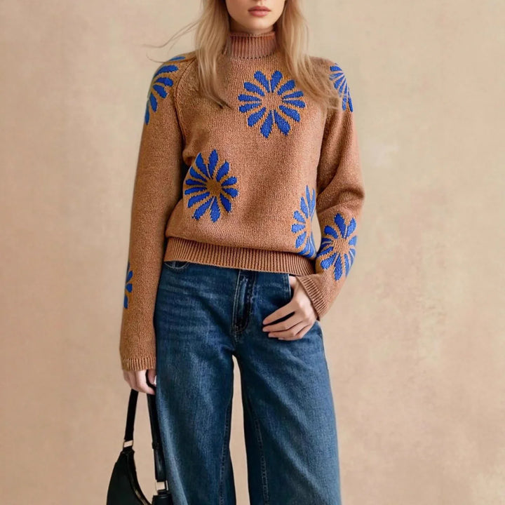 Woman wearing brown knit sweater with blue floral patterns and high-waisted blue jeans holding black handbag