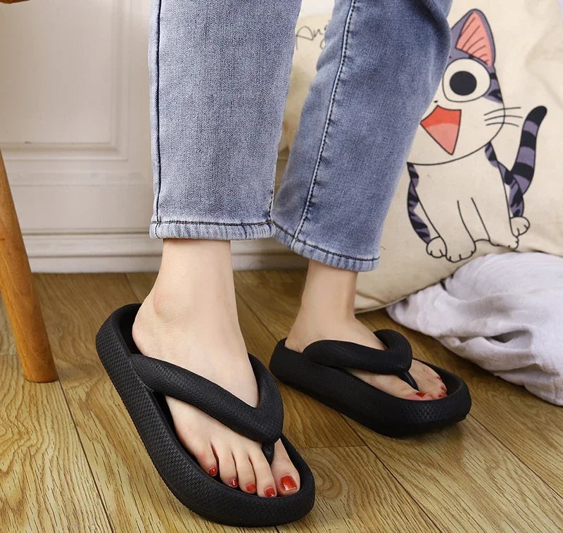 Person wearing black foam flip flop slippers on wooden floor with blue jeans and cat pillow