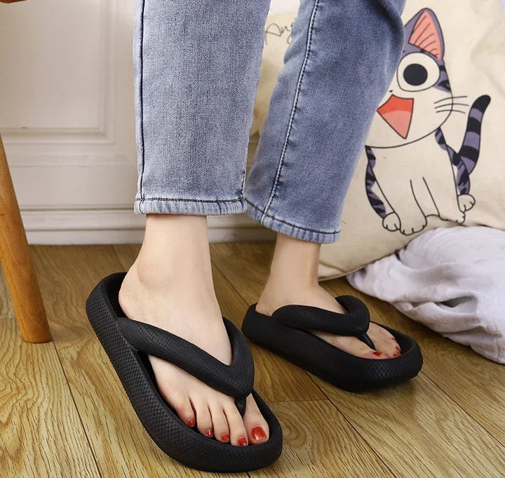 Person wearing black foam flip flop slippers on wooden floor with blue jeans and cat pillow