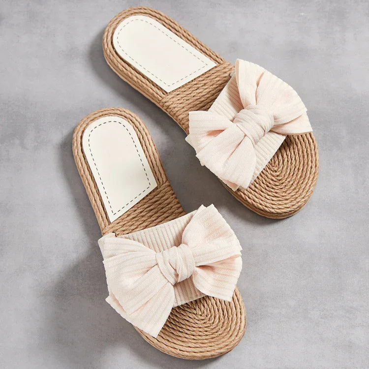 Beige slide sandals with large fabric bows on woven rope soles on gray background