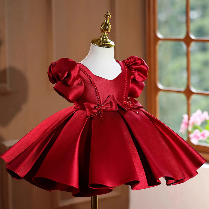 Elegant red satin baby dress with puff sleeves and decorative bows on mannequin near window