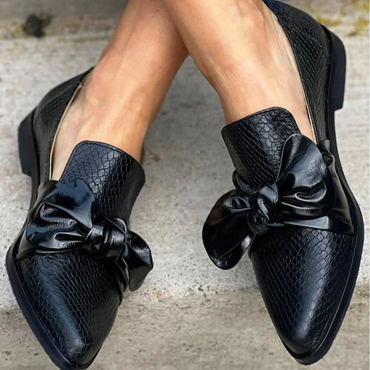 Black textured leather slip-on flats with large glossy bow detail on concrete floor