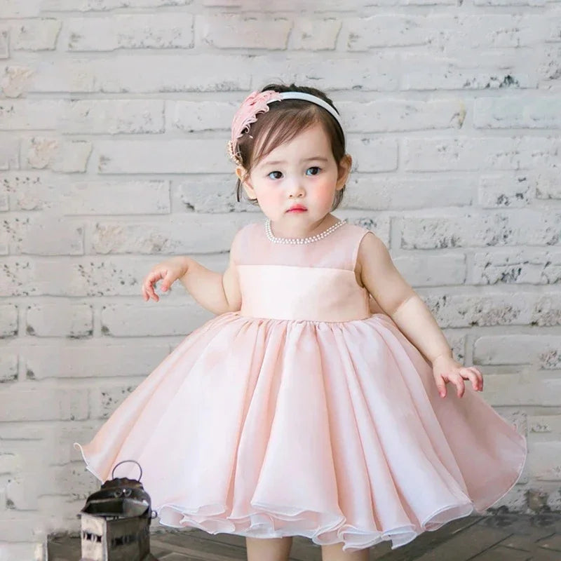 Toddler girl wearing a pink party dress and headband posing against a white brick wall