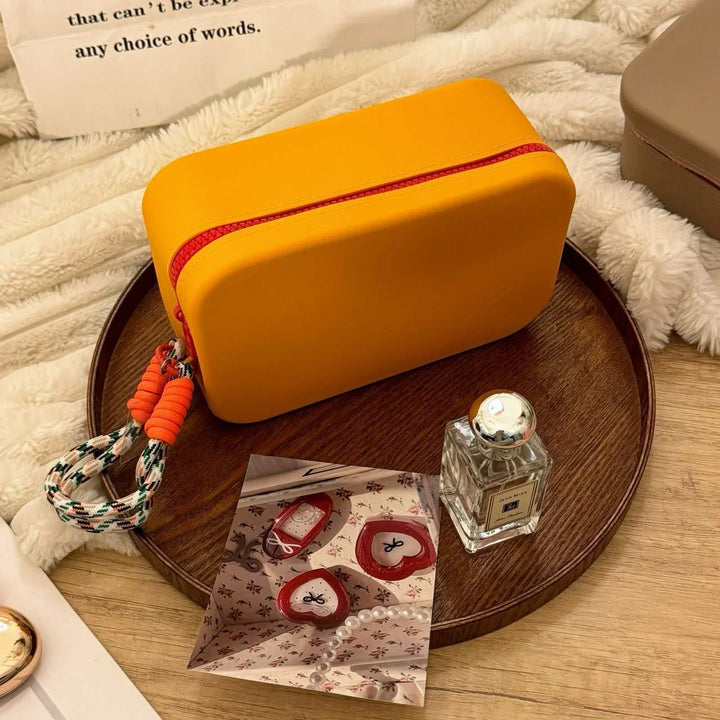 Yellow rectangular cosmetic bag with red zipper and wrist strap on wooden tray with perfume bottle and photo card