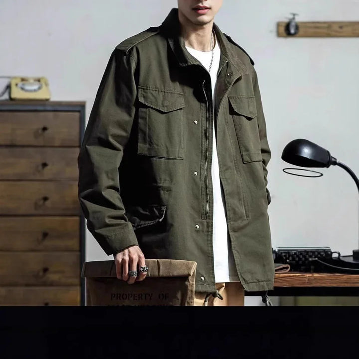 Man wearing olive green utility jacket over white shirt holding a brown clutch in vintage-style room