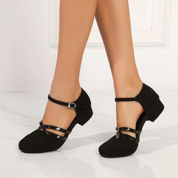 Black dance shoes with low heels and ankle straps worn on a white floor