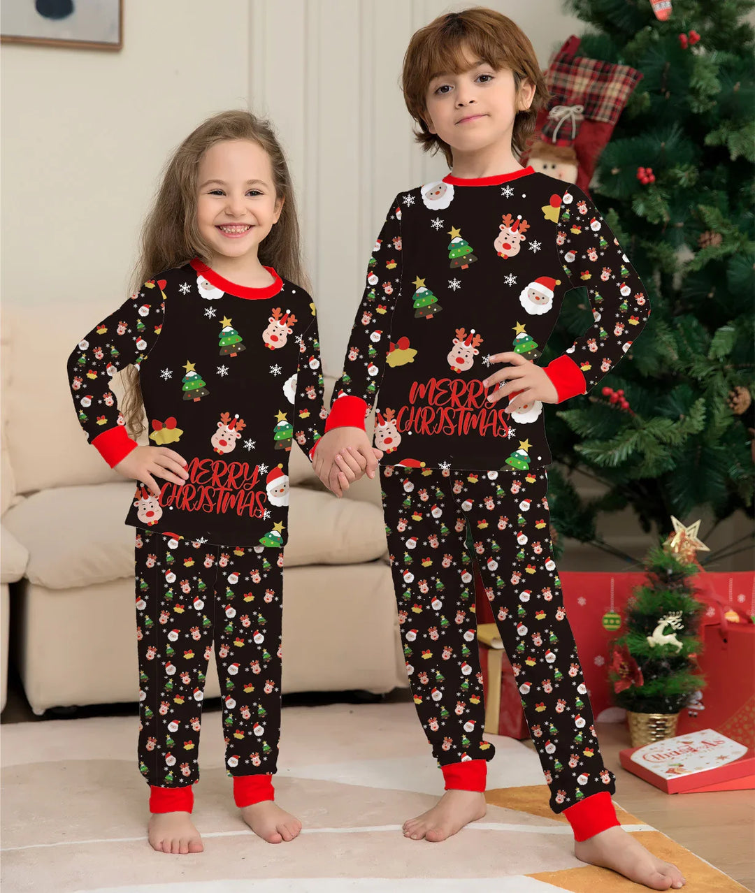 Two children in matching black Christmas pajamas with Santa, reindeer, and tree patterns, standing by a decorated tree