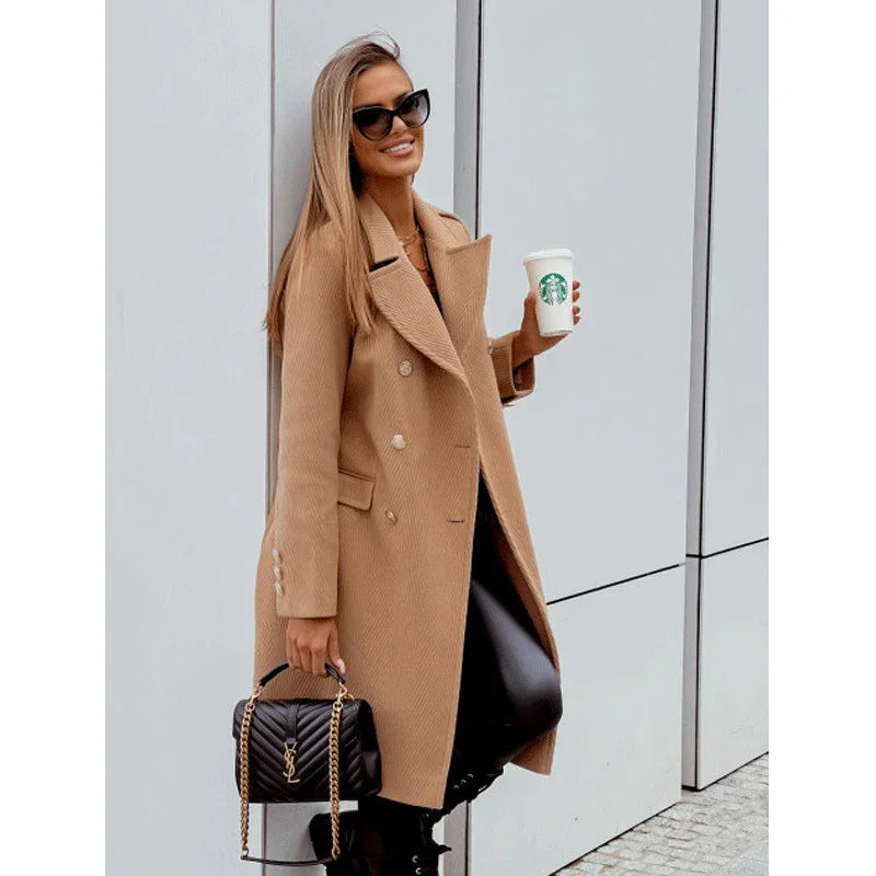 Stylish woman in tan double-breasted coat holding Starbucks cup and black designer handbag against modern wall