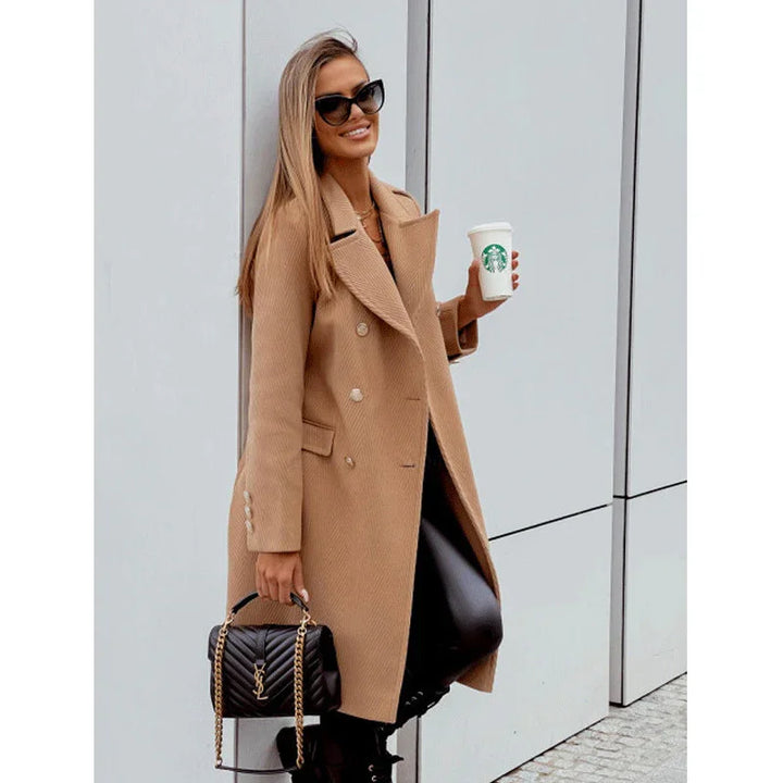 Stylish woman in tan double-breasted coat holding Starbucks cup and black designer handbag against modern wall