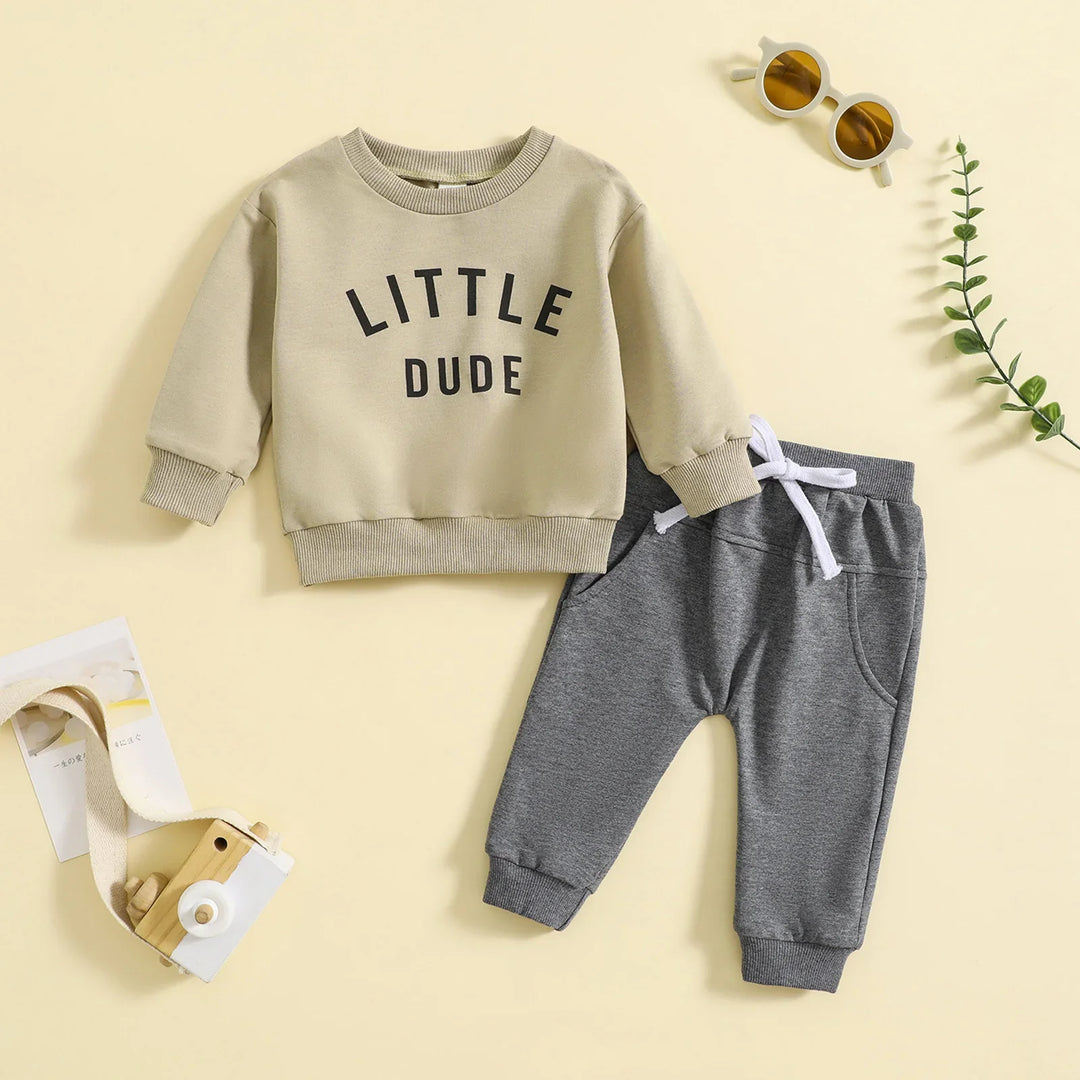Kids beige sweatshirt with Little Dude text and gray sweatpants set flat lay with sunglasses and toy camera
