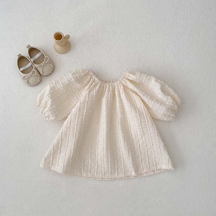Cream textured baby dress with puff sleeves, beige baby shoes, and small beige vase on white background