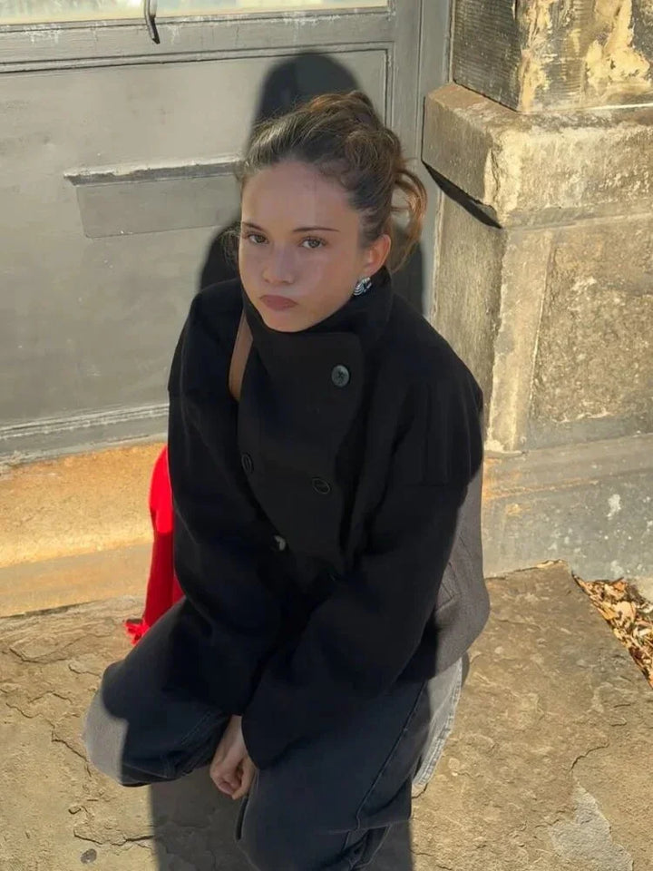 Young woman wearing black coat crouching by gray door and stone wall in sunlight