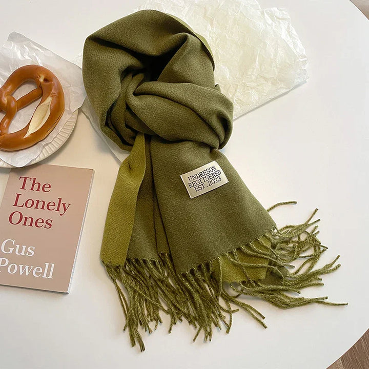 Olive green fringed scarf with Undreson Registered Est. 2023 label, book The Lonely Ones by Gus Powell, pretzel on a white plate