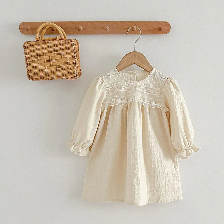 Cream-colored long sleeve baby dress with lace yoke on wooden hanger next to woven rattan handbag on wooden wall rack