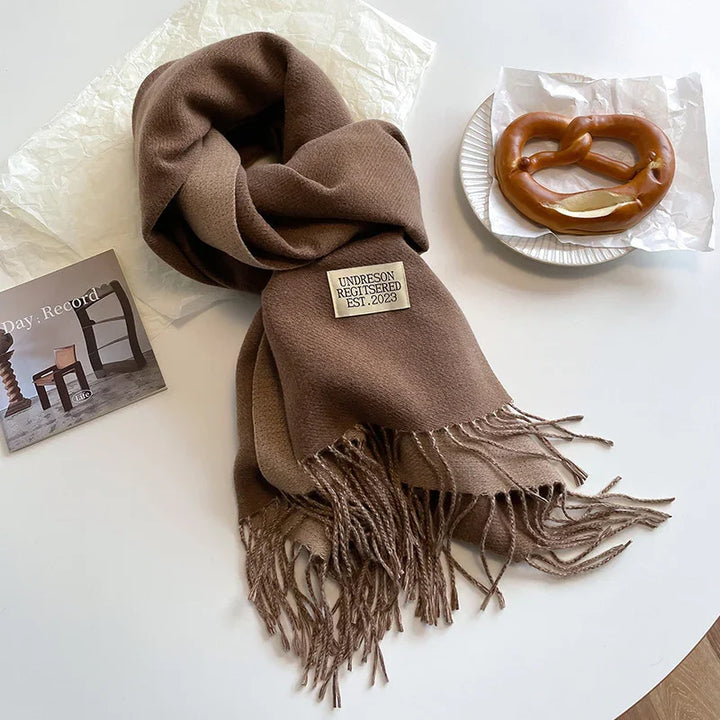 Brown fringed scarf labeled 'UNDRESON REGISTERED EST. 2023' on a white table with a pretzel on a plate