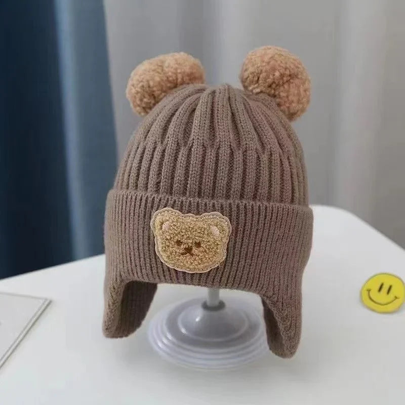 brown knitted baby hat with bear ears and a teddy bear patch on front, displayed on white stand