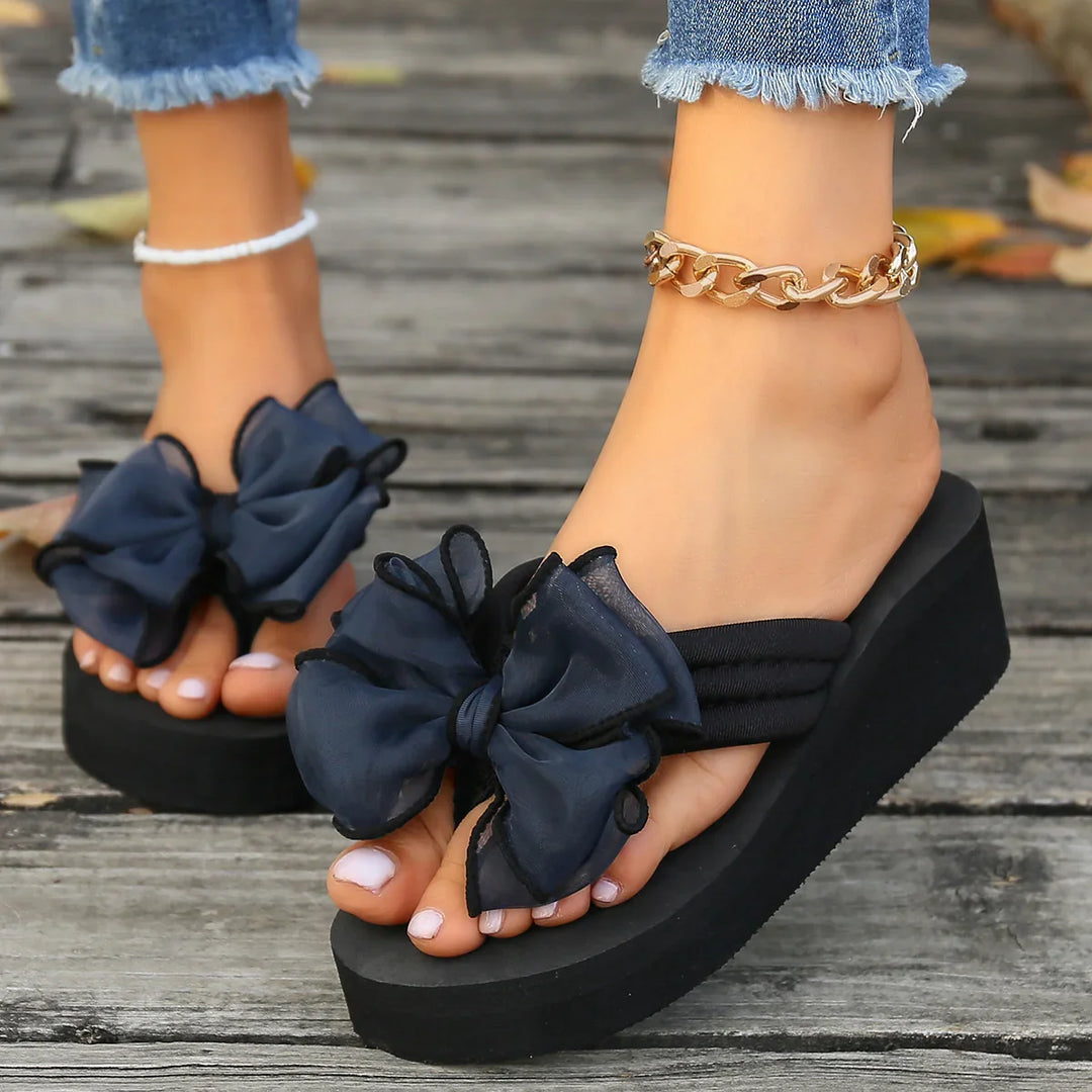 Close-up of feet in black platform sandals with large bows and gold chain anklet on wooden surface