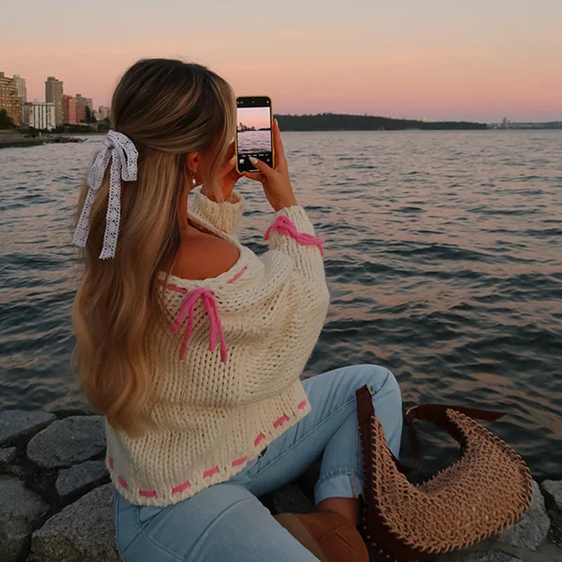 Woman in white knit sweater and jeans taking a photo of sunset over water by rocky shore