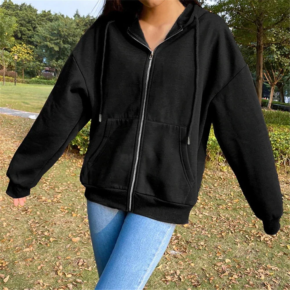 Person wearing black zip-up hoodie and blue jeans standing outdoors on grass with trees