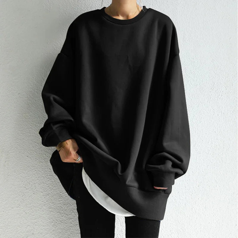 Person wearing oversized black sweatshirt with white shirt peek and black pants against gray wall
