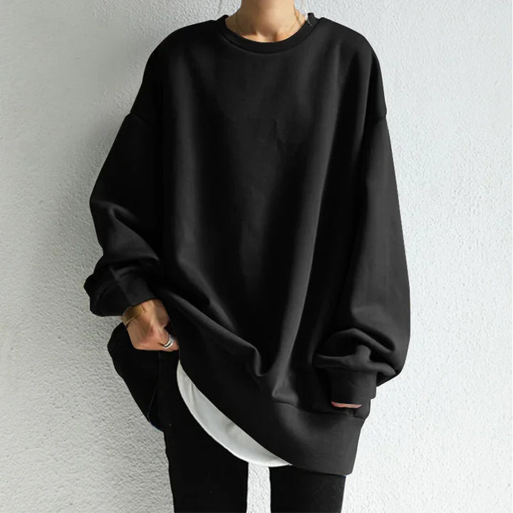 Person wearing oversized black sweatshirt with white shirt peek and black pants against gray wall