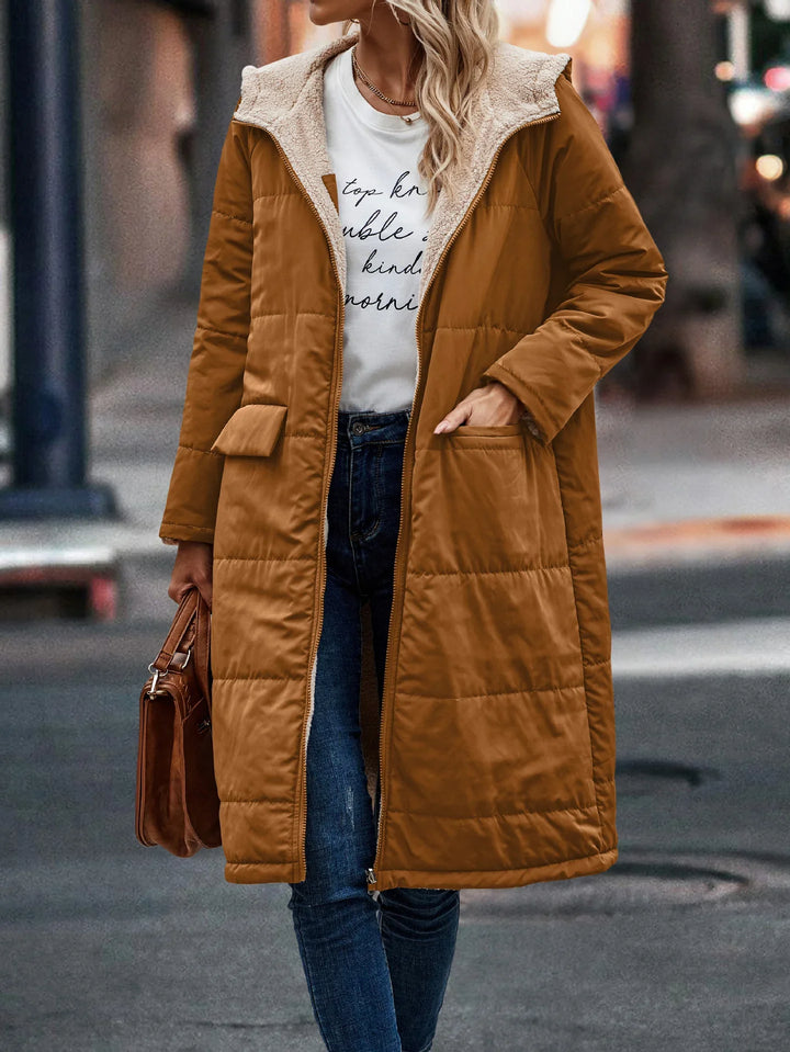 Woman wearing long brown puffer coat with faux shearling lining, white graphic tee, blue jeans, and carrying brown handbag