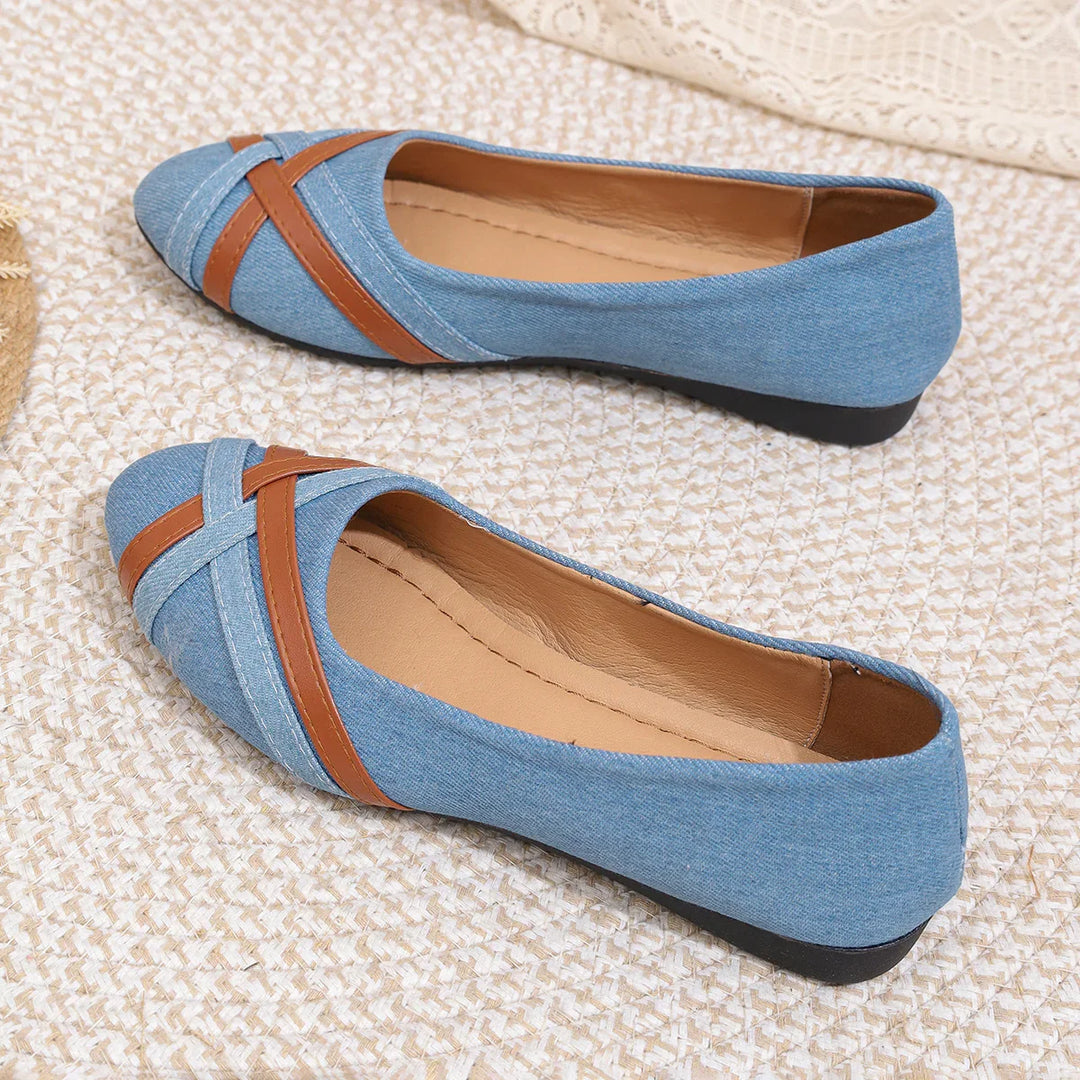 Blue denim flats with brown cross strap detail on textured beige surface