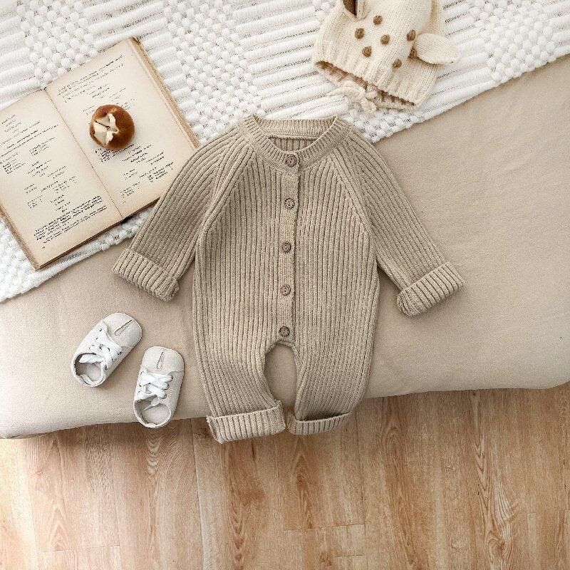 Beige knitted baby romper with buttons, white baby shoes, knitted animal hat, and open book on beige and white textured surface