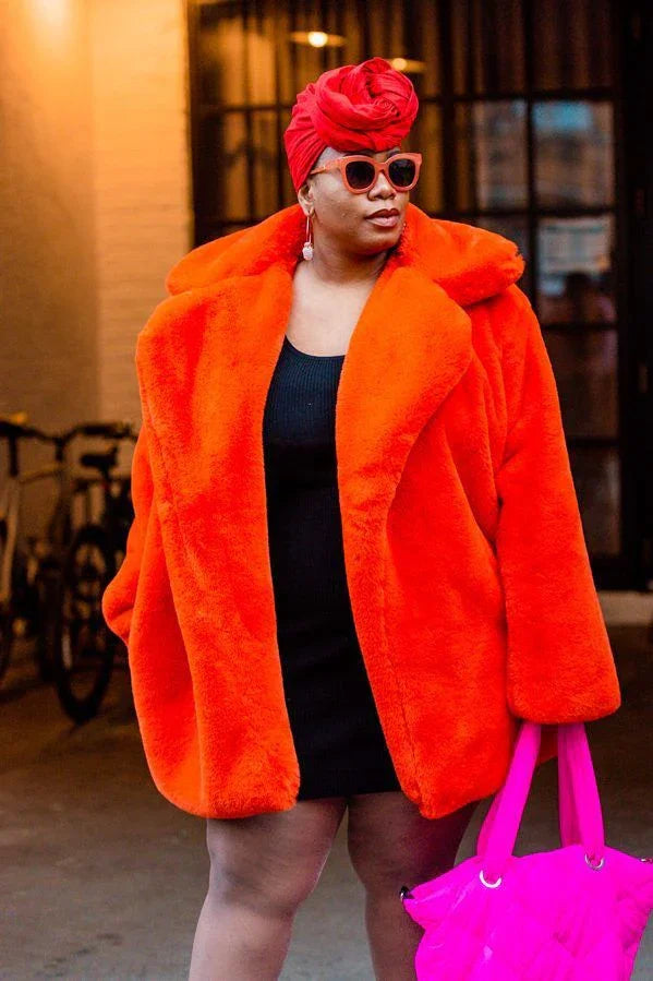 Woman wearing oversized bright orange fur coat, red turban, sunglasses, and holding a hot pink bag