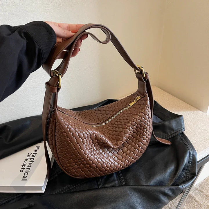 Hand holding brown woven leather shoulder bag with gold zipper over black leather jacket and book
