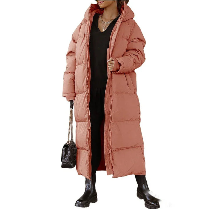 Woman wearing long dusty pink padded winter coat with hood, black boots, and carrying black handbag
