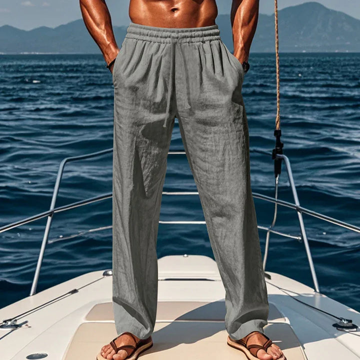 muscular man wearing gray linen pants and sandals standing on boat deck with ocean background