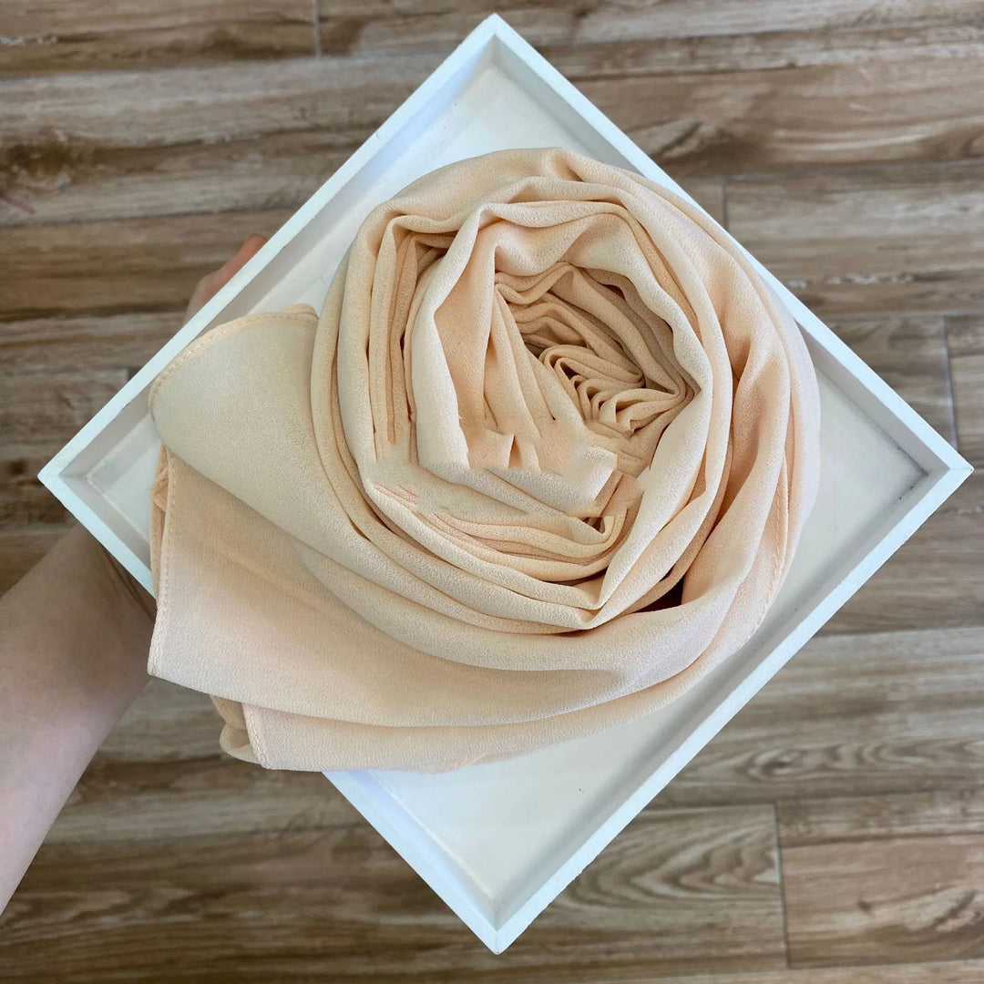 Beige fabric scarf neatly rolled inside a white square box held over wooden floor