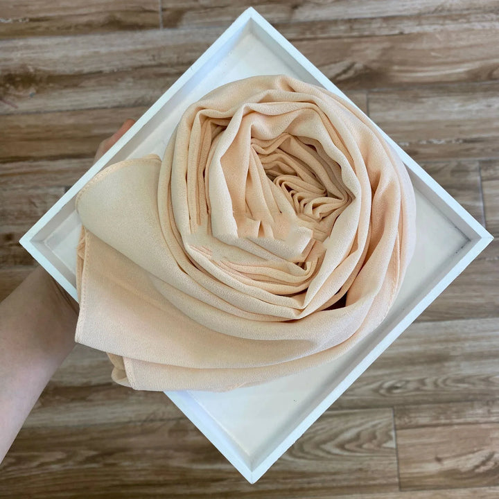 Beige fabric scarf neatly rolled inside a white square box held over wooden floor