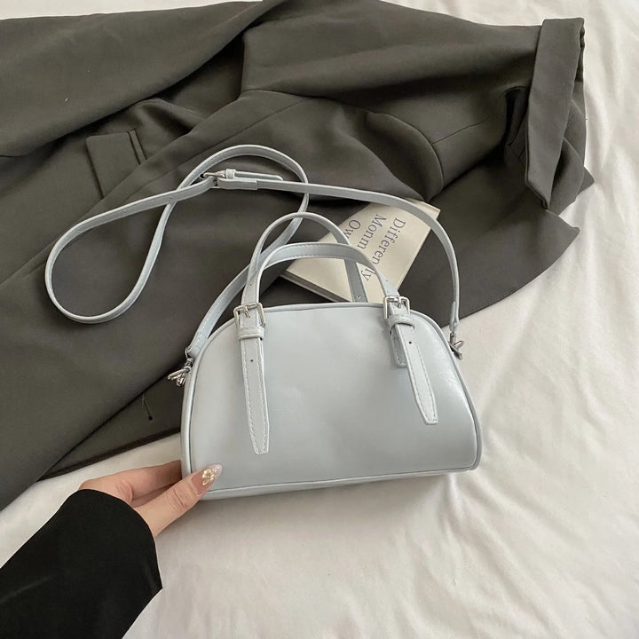 Light blue leather handbag with double buckled straps held over dark green jacket and book on white surface