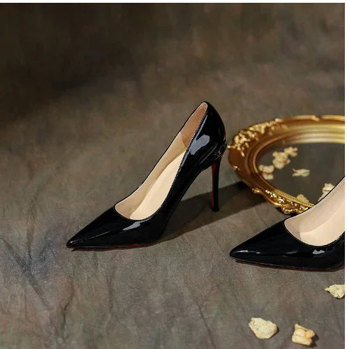 Black patent leather high heel pumps with pointed toes on gray surface near golden decorative mirror