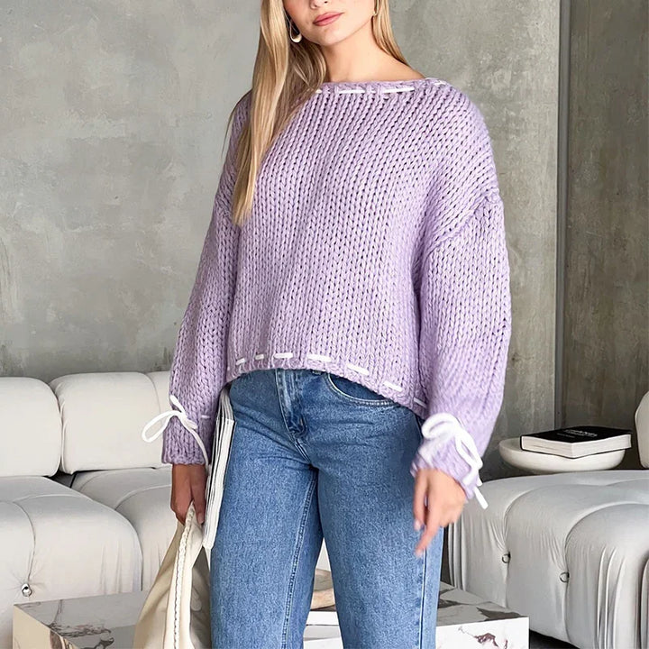 Woman wearing chunky lavender knit sweater with white ribbon details and blue jeans in modern living room